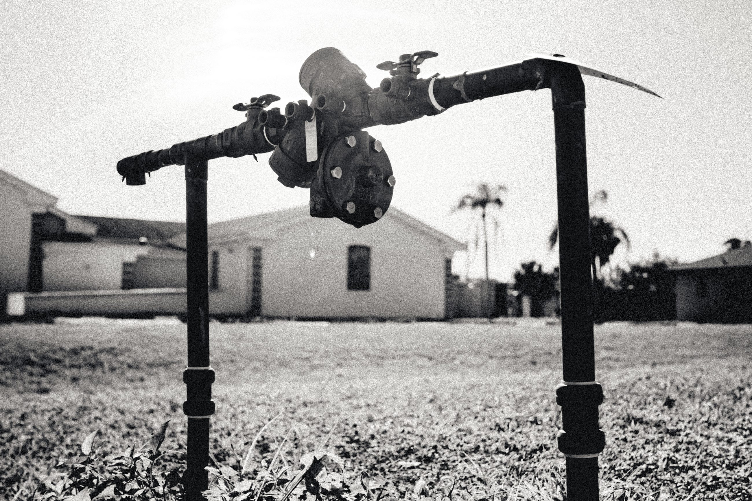 A wellhead of black pipe and valves standing waist-high on two legs in a mown yard, a low pale building and palms behind it.
