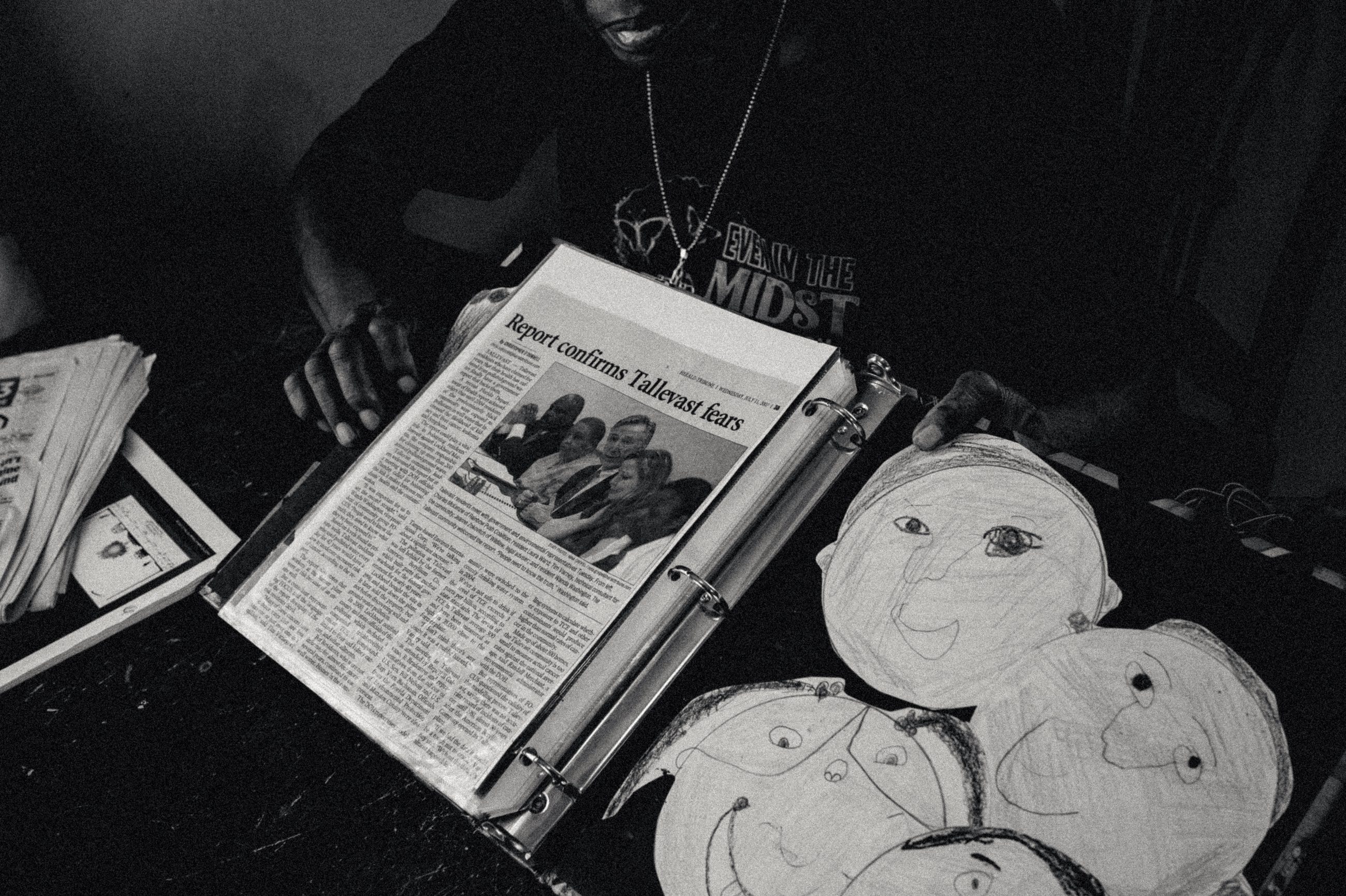 A person in a dark shirt holds open a ring binder of newspaper clippings, the top one headlined “Report confirms Tallevast fears.” Large crayon drawings of faces cover the opposite side of the open binder.